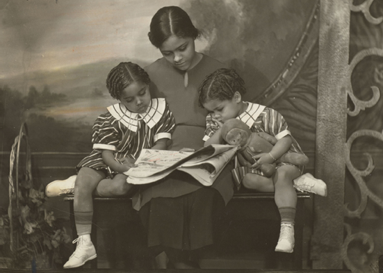 A woman sitting on a bench reads from a newspaper to two small children who are leaning against her.