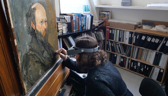An art conservator wearing a magnifier headset does detailed work on a corner of a painting, Paul Cézanne’s Self-Portrait (1878-80).